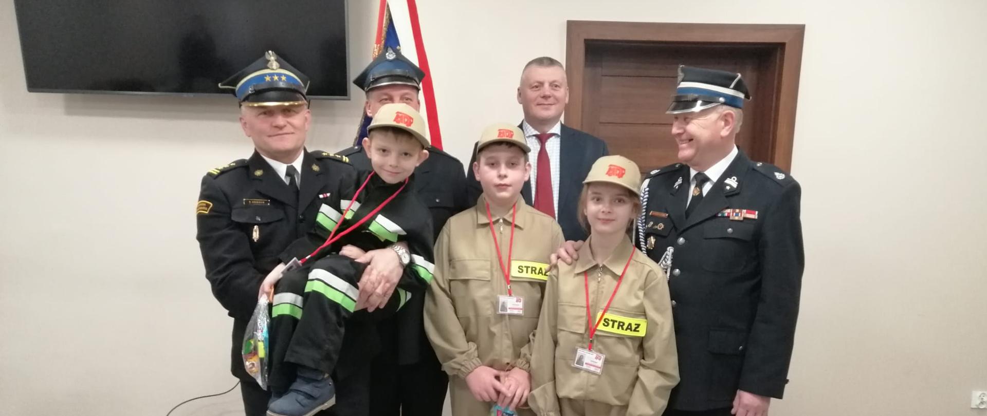 Na zdjęciu widać pozujących 4 mężczyzn do zdjęcia oraz 3 dzieciaczki w mundurach z młodzieżowej drużyny pożarniczej MDP Witosławice. Na zdjęciu jeden z mężczyzn to Komendant Powiatowy PSP w Ostrowcu Św., który na swoich rękach trzyma jedno z dzieci. Wszyscy pozujący są uśmiechnięci. Zdjęcie wykonane w remizie OSP Witosławice.