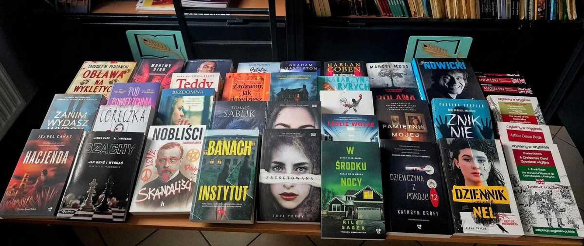 Na zdjęciu znajduje się regał biblioteczny wypełniony książkami oraz stół, na którym wyłożono dużą liczbę różnych tytułów literackich.