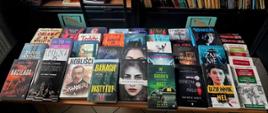 Na zdjęciu znajduje się regał biblioteczny wypełniony książkami oraz stół, na którym wyłożono dużą liczbę różnych tytułów literackich.