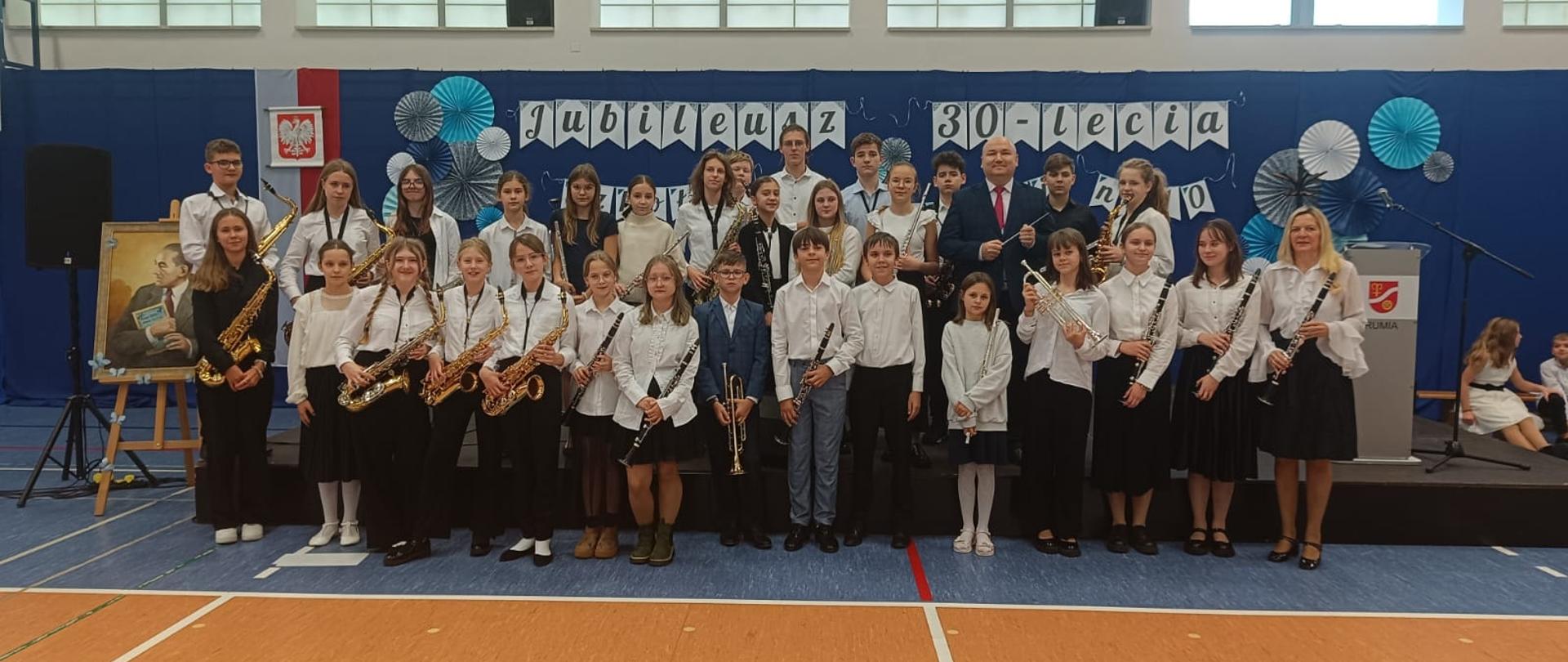 Orkiestra dęta pozuje do zdjęcia w strojach galowych na sali gimnastycznej SP nr 10 w Rumi. Wszystkie osoby stoją trzymając instrumenty.