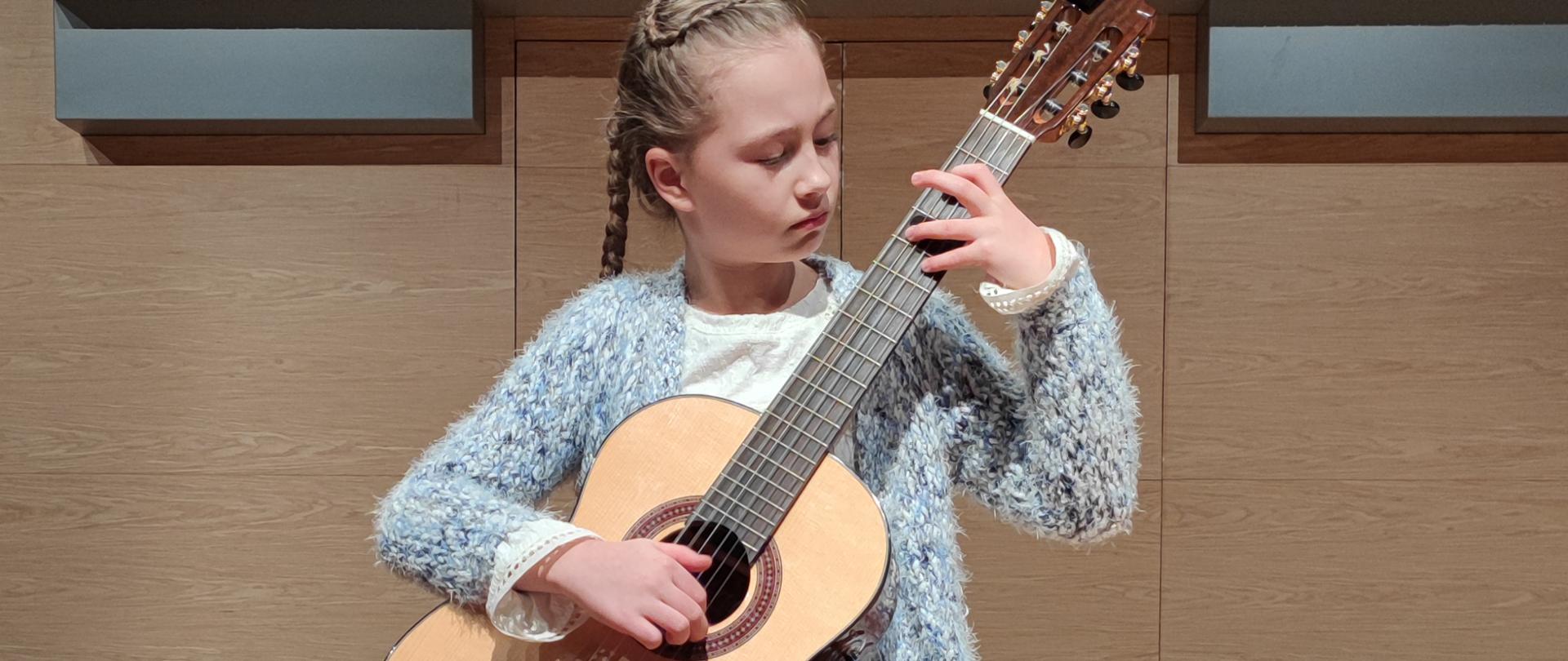 Dziewczynka w jasnym swetrze i ciemnych spodniach trzyma w rękach gitarę klasyczną. Głowę ma skierowaną w lewą stronę i patrzę na palce lewej ręki, które grają na gryfie gitary. W tle na ścianie jest namalowana klawiatura fortepianu, nad którą znajduje się duża ilość piszczałek organowych srebrnego koloru o różnej długości.