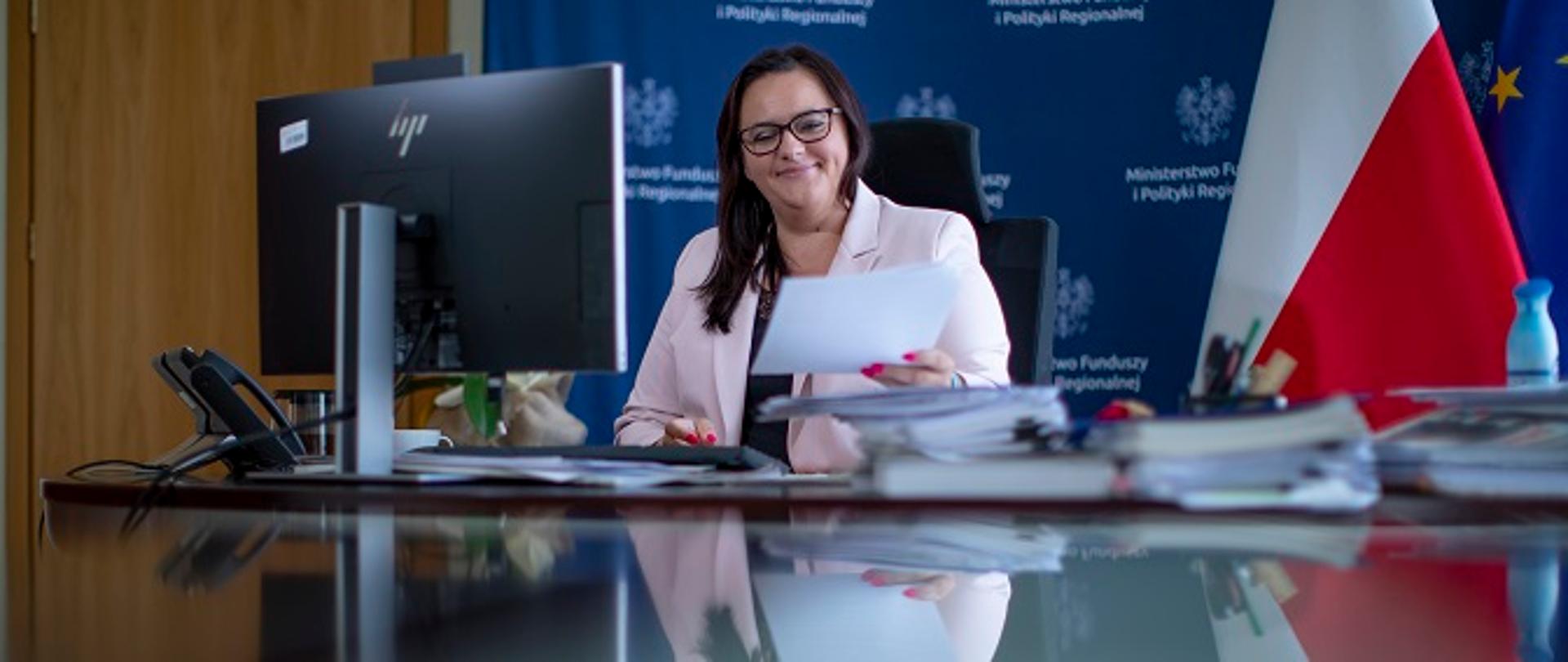 Na zdjęciu wiceminister funduszy i polityki regionalnej Małgorzata Jarosińska-Jedynak siedząca przy biurku i trzymająca w ręce dokumenty