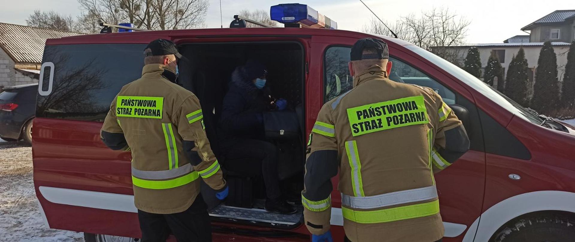Dwóch strażaków odwróconych tyłem przed samochodem. W środku osoba w maseczce. Przewóz na szczepienia. 
