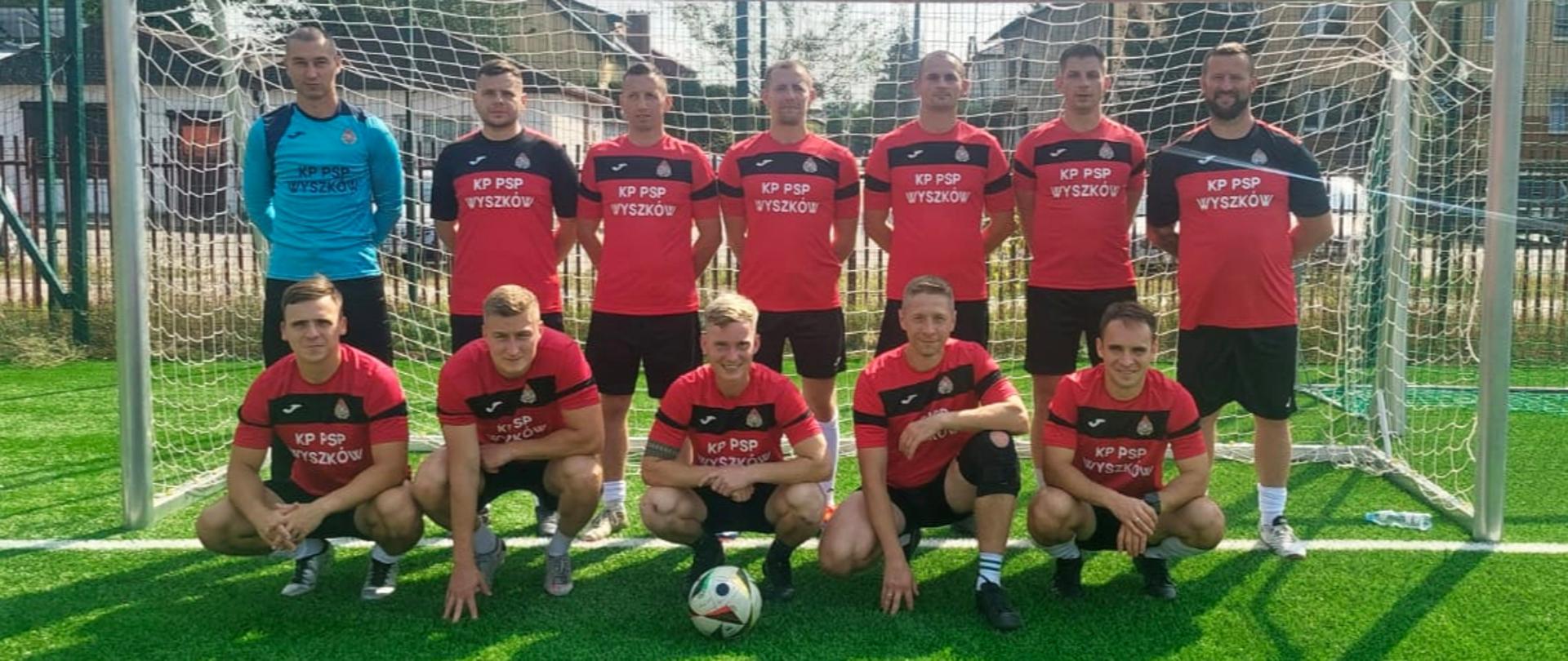 Grupowe zdjęcie strażaków w czerwono-czarnych strojach sportowych na tle bramki.