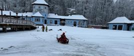 Strażacy w czerwonych ubraniach wypornościowych do pracy w wodzie ćwiczą podejmowanie osoby poszkodowanej, pod którą załamał się lód i wpadła do wody. Na zdjęciu strażacy na saniach lodowych. Zamarznięte jezioro w zimowej aurze. Strażacy wykonują manewr sciągania sań lodowych na brzeg jeziora.