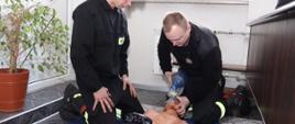 na podłodze leży fantom do nauki resuscytacji krążeniowo-oddechowej przy którym klęczą dwaj strażacy osp ubrani w czarne ubranie koszarowe jeden trzyma manekina za głowę i przykłada mu do twarzy worek ambu a drugi przygląda się resuscytacji