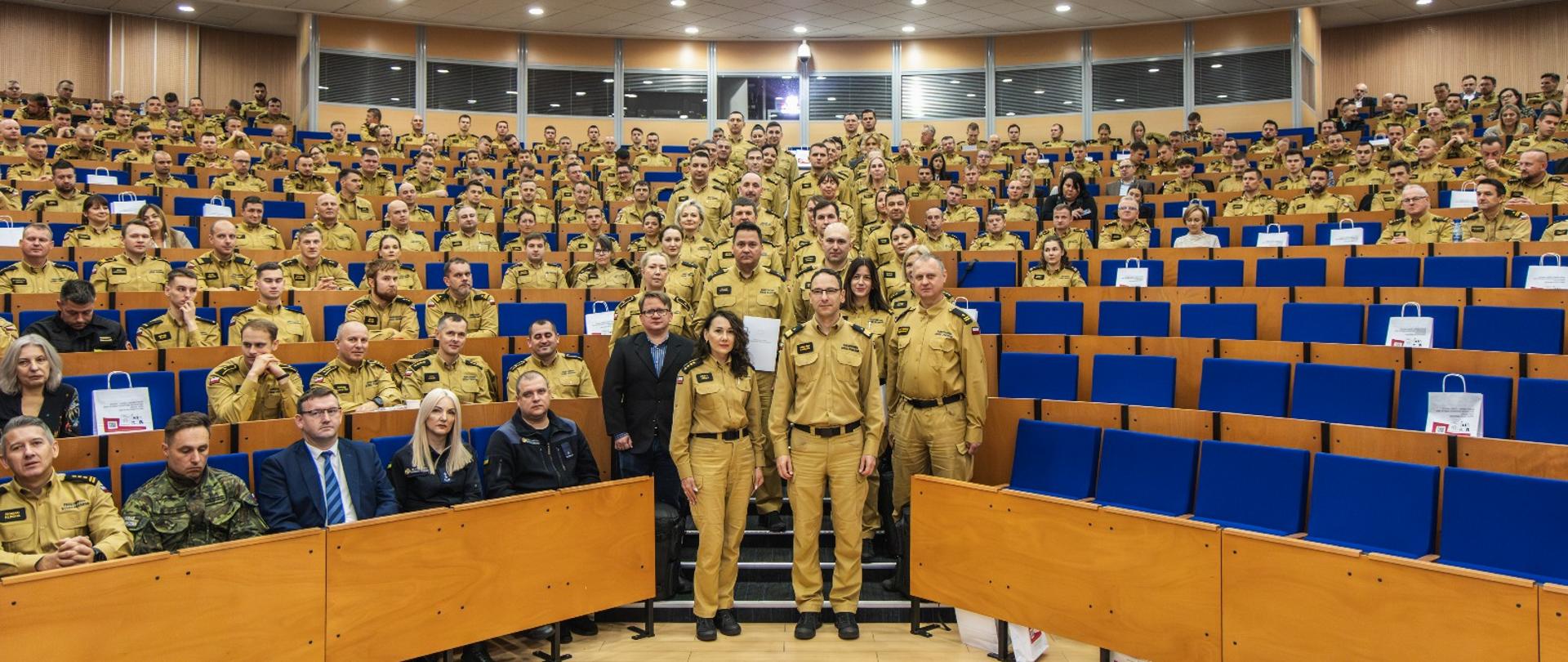 Zdjęcie grupowe uczestników konferencji PSP w auli Centralnej Szkoły PSP w Częstochowie. Wszyscy stoją i siedzą w mundurach, patrząc w stronę obiektywu.