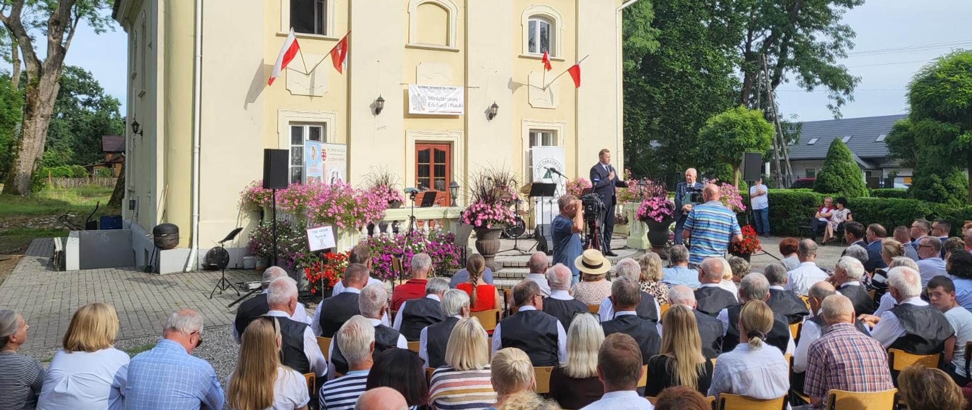 Zdjęcie z tyłu, na dworze przed żółtym budynkiem ustawionych w rzędy krzesłach siedzą ludzie, przed nimi mężczyzna w garniturze mówi do mikrofonu.