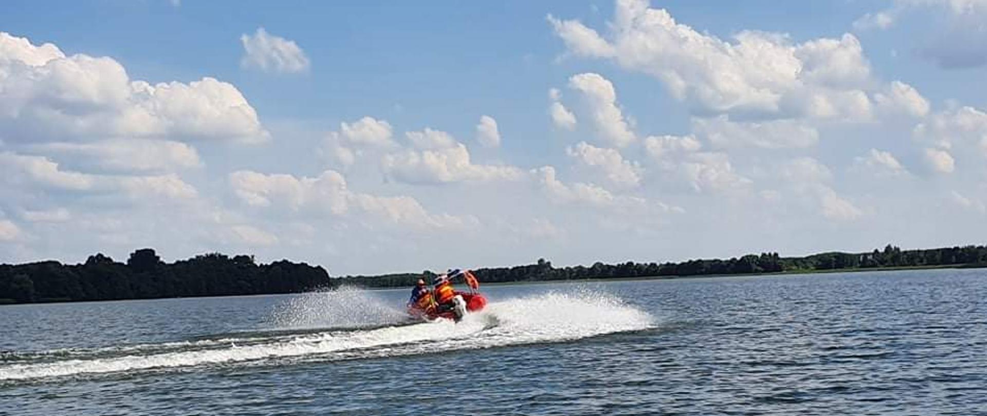 Zdjęcie wykonano porą dzienną, na zdjęciu łódź motorowa Państwowej Straży Pożarnej z dwoma strażakami, funkcjonariuszem komendy policji i ratownikiem wodnym na pokładzie, płyną po zbiorniku wodnym. 