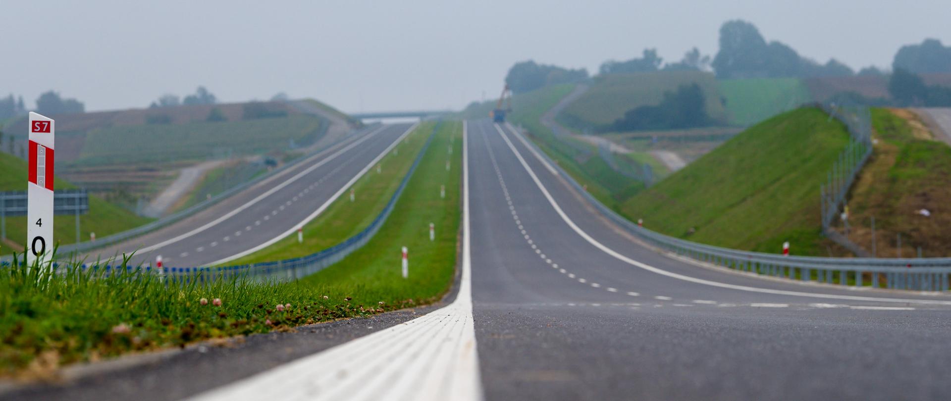 Droga ekspresowa S7 na odcinku Szczepanowice - Widoma tuż przed oddaniem do ruchu. Dwujezdniowa droga ekspresowa, po dwa pasy ruchu w obu kierunkach. Zdjęcie wykonane blisko gruntu. Widoczny biały pas oznakowania oddzielający pas ruchu od krawędzi drogi. Po lewej biały słupek pikietażowy z nr drogi ekspresowej.