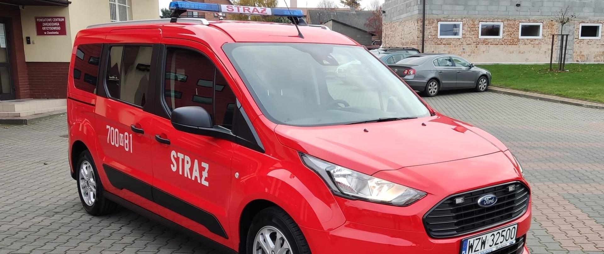 Lekki samochód kwatermistrzowski na podwoziu Ford Tourneo Connect w kolorze czerwonym. Zdjęcie zostało zrobione na placu Komendy Powiatowej Państwowej Straży Pożarnej w Zwoleniu. W tle widać wejście do głównego budynku Komendy, drugi budynek oraz samochody zaparkowane na parkingu.