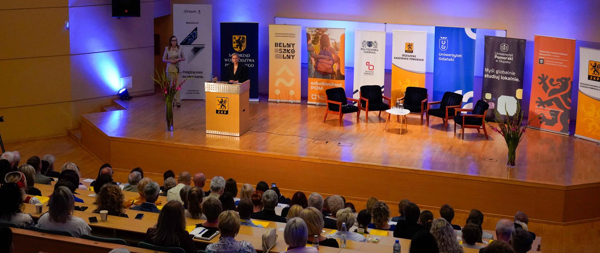 sala konferencyjna, z rzędami krzeseł dla publiczności. Na scenie znajduje się mównica, za którą stoi kobieta. W tle widać banery w różnych kolorach z napisami „BELNY SZKOLNY” i logo Uniwersytetu Gdańskiego. Na dużym ekranie nad sceną widnieje napis „KONGRES SZKOLNYCH KASZËBSCZÉGÒ JÃZËKA Gdańsk, 13 sewnika 2025 roku” oraz nazwisko prelegentki „BARBARA NOWACKA Ministrõ Norõdny Edukacje”.