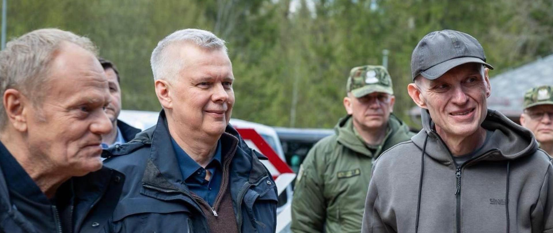 Na pierwszym planie trzy osoby - od prawej Andrzej Poczobut, minister Tomasz Siemoniak i premier Donald Tusk. Za Andrzejem Poczobutem stoi dwóch funkcjonariuszy Straży Granicznej. W tle widać samochody i drzewa.