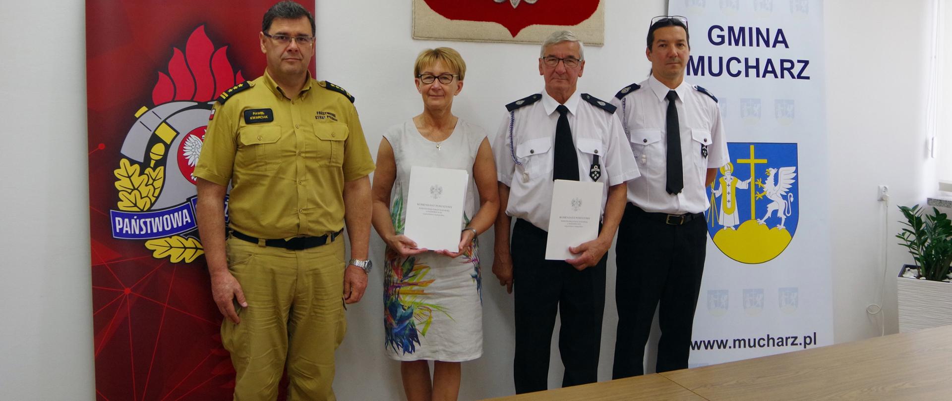 Widok z przodu. Uczestnicy spotkania stoją za stołem na tle godła Polski i rolapów KP PSP Wadowice i Gminy Mucharz. Od lewej: komendant powiatowy PSP, wójt gminy, prezes OSP i naczelnik OSP.