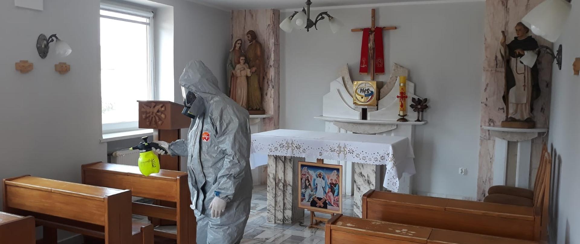 Strażak w odpowiednim stroju ochronnym za pomocą opryskiwacza ręcznego dokonuje dezynfekcji pomieszczenia, w którym znajduje się kaplica kościelna. Po obu stronach są drewniane ławki a w centrum znajduje się ołtarz. 