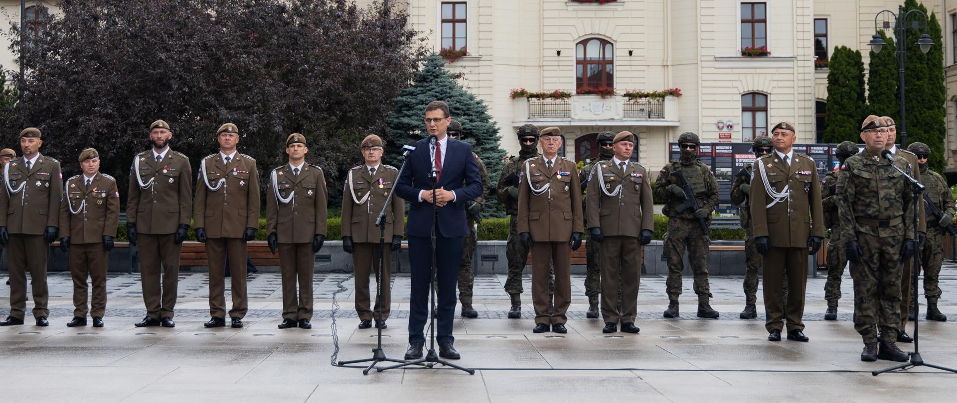 Wojewoda Kujawsko-Pomorski - Michał Sztybel - przemawia na tle bydgoskiego ratusza na Starym Runku w Bydgoszczy. Na drugim planie stoją żołnierze w mundurach galowych. 