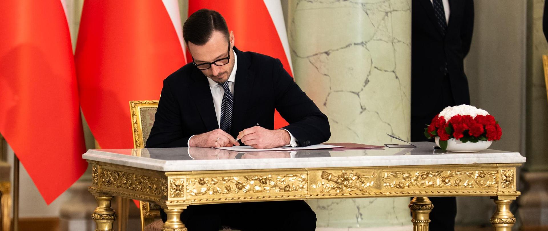 Zaprzysiężenie nowego Ministra Aktywów Państwowych Jakuba Jaworowskiego. Minister siedzi przy stole i podpisuje rotę przysięgi.