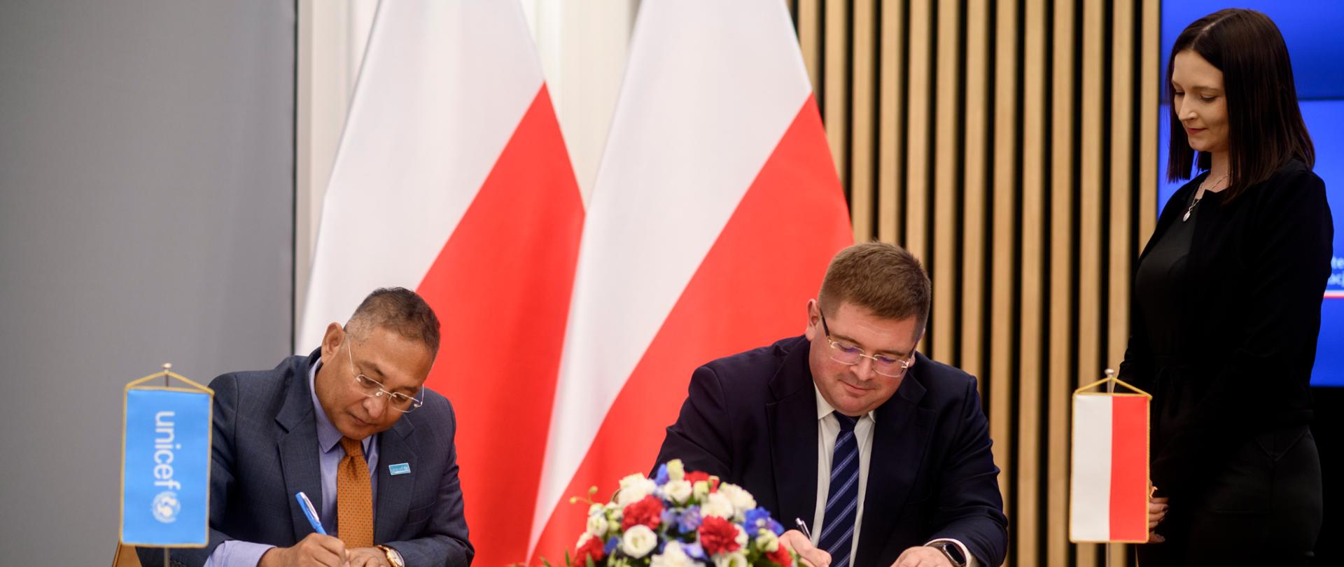 Przy stoliku siedzi wiceminister Rzymkowski i mężczyzna w garniturze, obaj podpisują dokumenty, nad nimi stoi kobieta w czarnym ubraniu, za nimi kilka polskich flag.