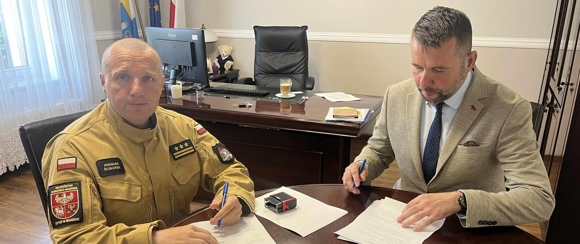 Po prawej stronie siedzi mężczyzna w garniturze,. Po lewej stronie siedzi strażak w piaskowym mundurze z emblematami i stopniami służbowymi. Zdjęcie dokumentuje ważny moment – prawdopodobnie podpisanie umowy, porozumienia lub wręczenie nominacji związanej z działalnością strażacką. Atmosfera jest oficjalna, ale przyjazna.