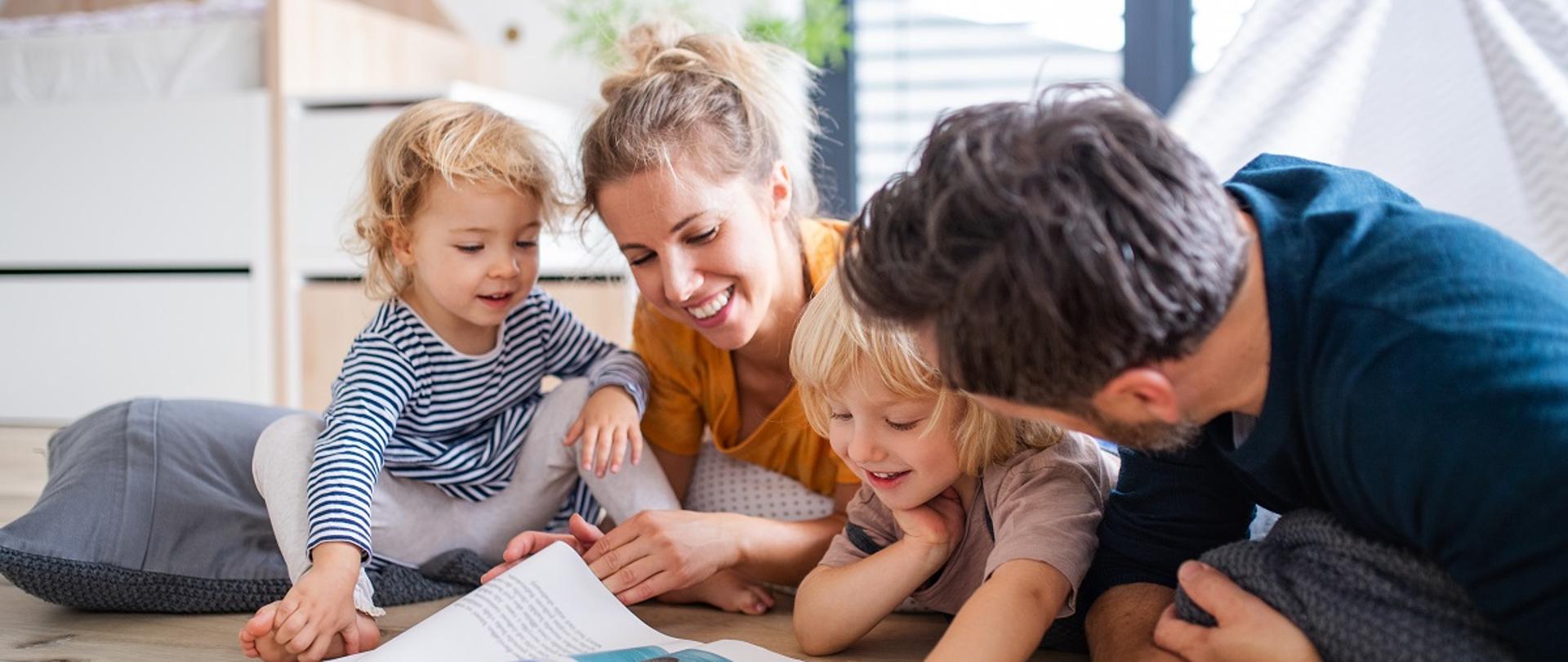 Front view of young family with two small children indoors in bedroom reading a book.