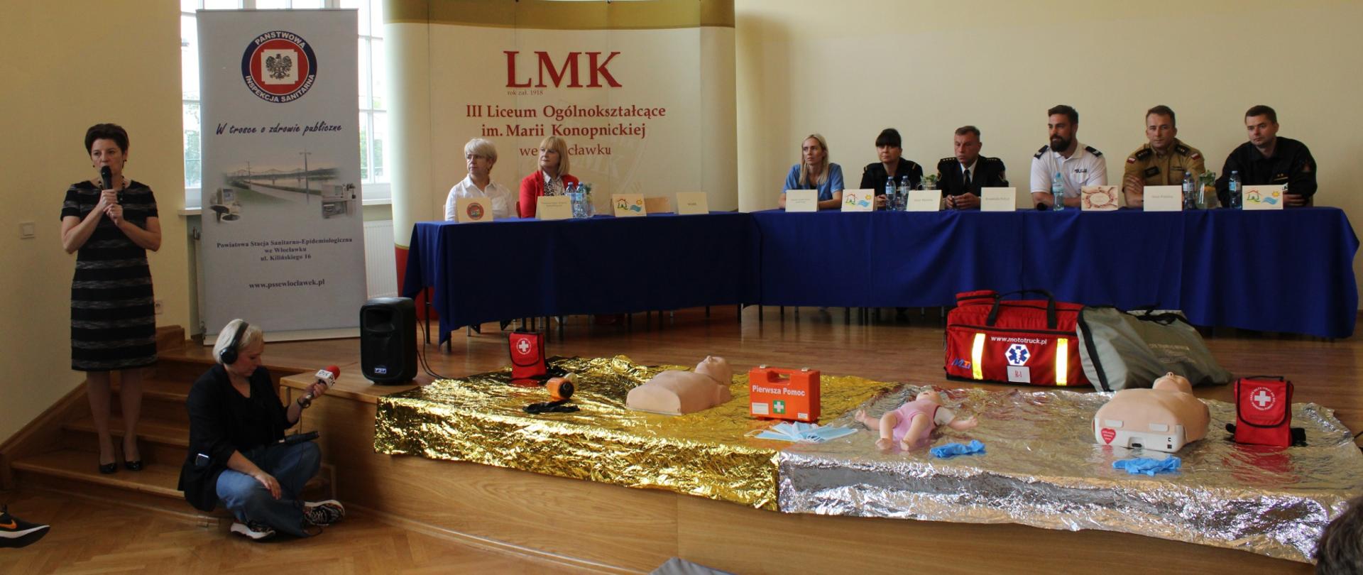 stół na scenie zasiadają za nim osoby powiązane z bezpieczeństwem zdrowia i obywateli