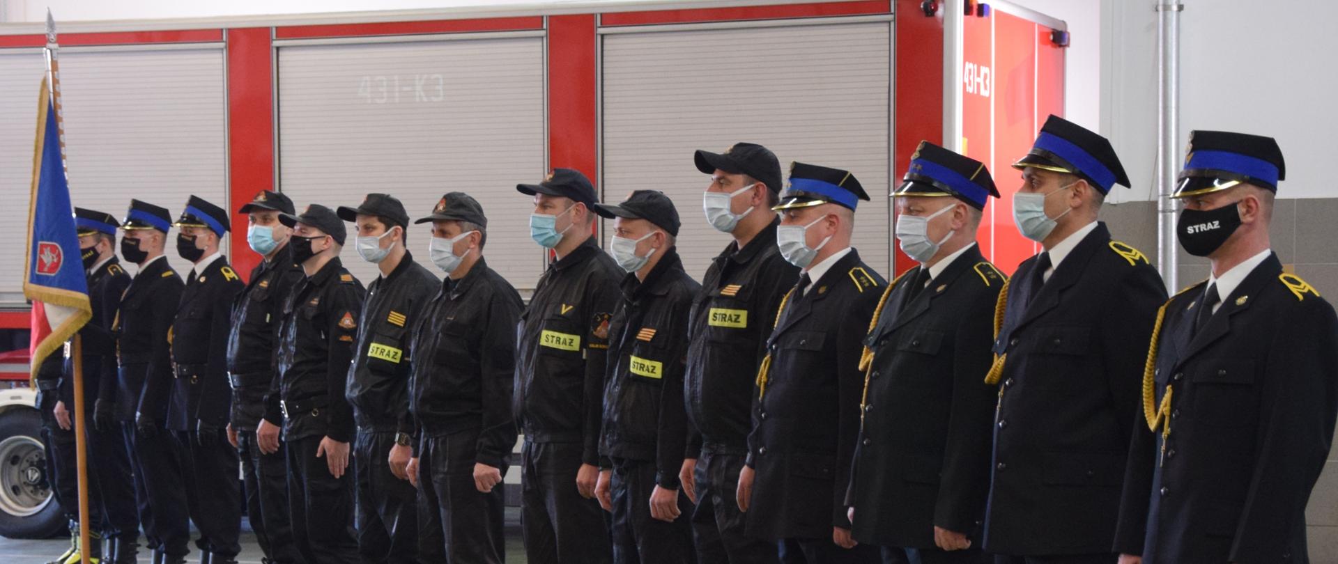 Na zdjęciu widać ustawionych w szeregu strażaków z maseczkami ochronnymi ma twarzach. Część z nich jest w umundurowaniu galowym a pozostali w stroju koszarowym, w tle po lewej stronie widać sztandar.