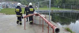 Akcja na składowisku odpadów. Przeciwpożarowy zbiornik wody. Przy nim trzech strażaków. W tle pryzma odpadów, betonowa ściana i drzewa.