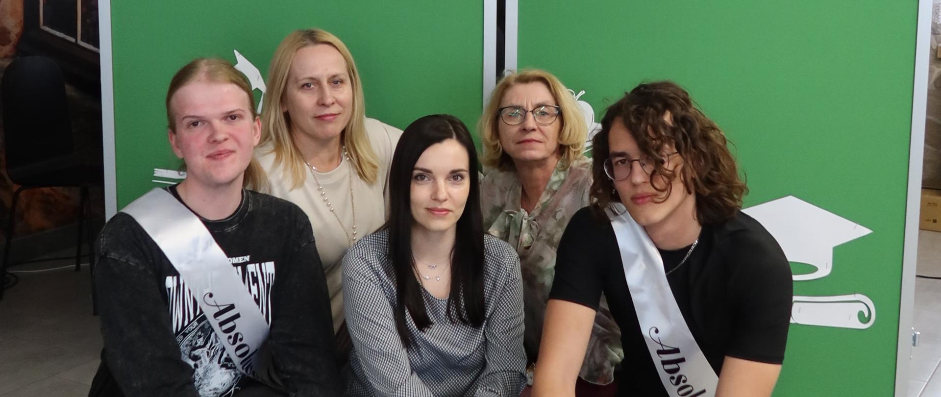 Dwaj absolwenci i trzy nauczycielki pozują do zdjęcia na tle tablicy z napisem "Do zobaczenia, drodzy Absolwenci".