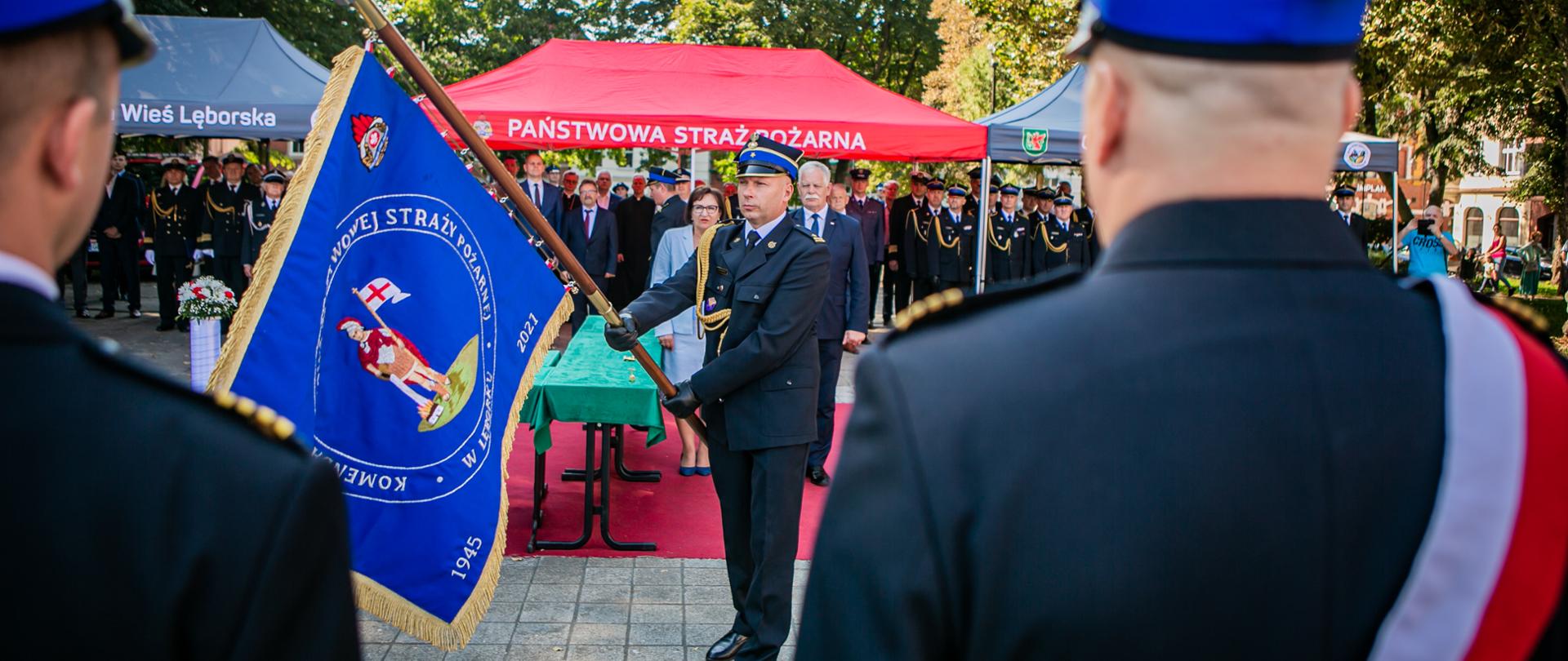 Komendant Powiatowy Państwowej Straży Pożarnej w Lęborku młodszy brygadier Piotr Krzemiński dokonuje prezentacji sztandaru. Na drugiej stronie płata sztandaru znajduje się wizerunek św. Floriana, a wokół niego napis „Komenda Powiatowa Państwowej Straży Pożarnej w Lęborku”. Na sztandarze umieszczone są również: herb powiatu lęborskiego, logo Państwowej Straży Pożarnej oraz daty: 1945 – rok powołania Okręgowego Pogotowia Straży Pożarnej w Lęborku, 2021 – rok nadania sztandaru Komendzie Powiatowej PSP w Lęborku. Poczet sztandarowy stoi przed komendantem. W tle widoczni wręczający sztandar Starosta Lęborki Pani Alicja Zajączkowska Przewodniczący Społecznego Komitetu Fundacji Sztandaru oraz Wicewojewoda Pomorski Pan Mariusz Łuczyk. Za nimi stoją goście zgromadzeni na uroczystości. 