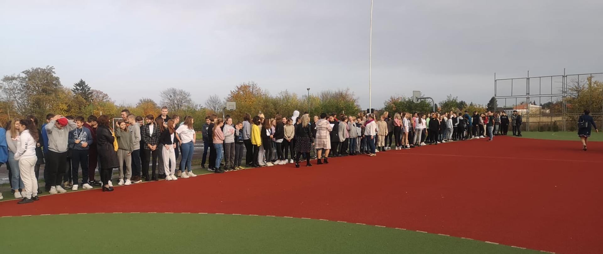 Zdjęcie przedstawia próbną ewakuacją Publicznej Szkole Podstawowej nr 4im Osadników Wojskowych w Świdwinie. Na boisku znajdują się ewakuowane osoby. 