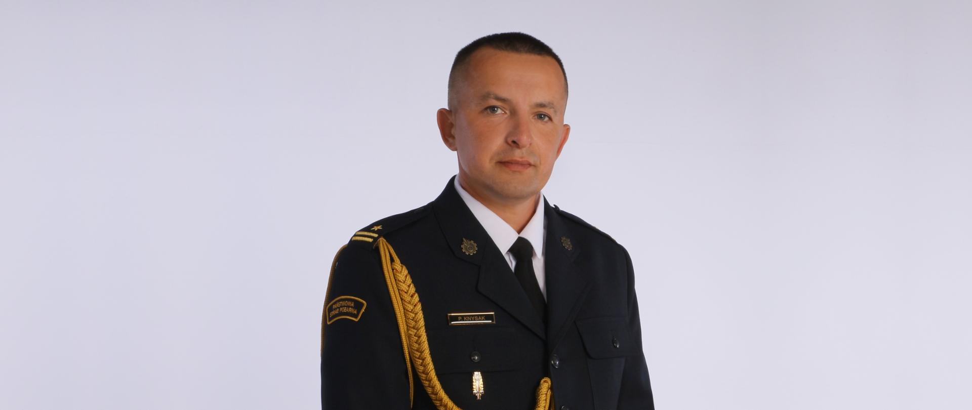 Powyższe zdjęcie to portret mężczyzny w ciemnym, galowym mundurze, prawdopodobnie oficera służb mundurowych.
Mężczyzna ma krótkie, starannie ułożone, jasne włosy i patrzy lekko w bok od obiektywu. Jego twarz jest poważna. Ubrany jest w ciemny mundur z białą koszulą i czarnym krawatem.
Na jego prawym ramieniu widać naszywkę z napisem "PAŃSTWOWA STRAŻ POŻARNA", co sugeruje przynależność do tej formacji. Na lewym ramieniu widoczny jest pagon ze złotą gwiazdką i dwiema belkami (co sugeruje stopień oficerski) oraz ozdobny, złoty pleciony sznur. Na piersi, po lewej stronie, przypięta jest złota plakietka z napisem "P. KNYSAK".