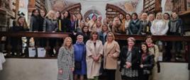 Zdjęcie grupowe w Bibliotece Poaugustyńskiej w Żaganiu. Grupa uśmiechniętych kobiet, uczestniczek konferencji, stojących na tle historycznego, pięknie zdobionego wnętrza Biblioteki Poaugustyńskiej w Żaganiu. Za nimi widać regały z książkami oraz freski na suficie przedstawiające barwne sceny historyczne.