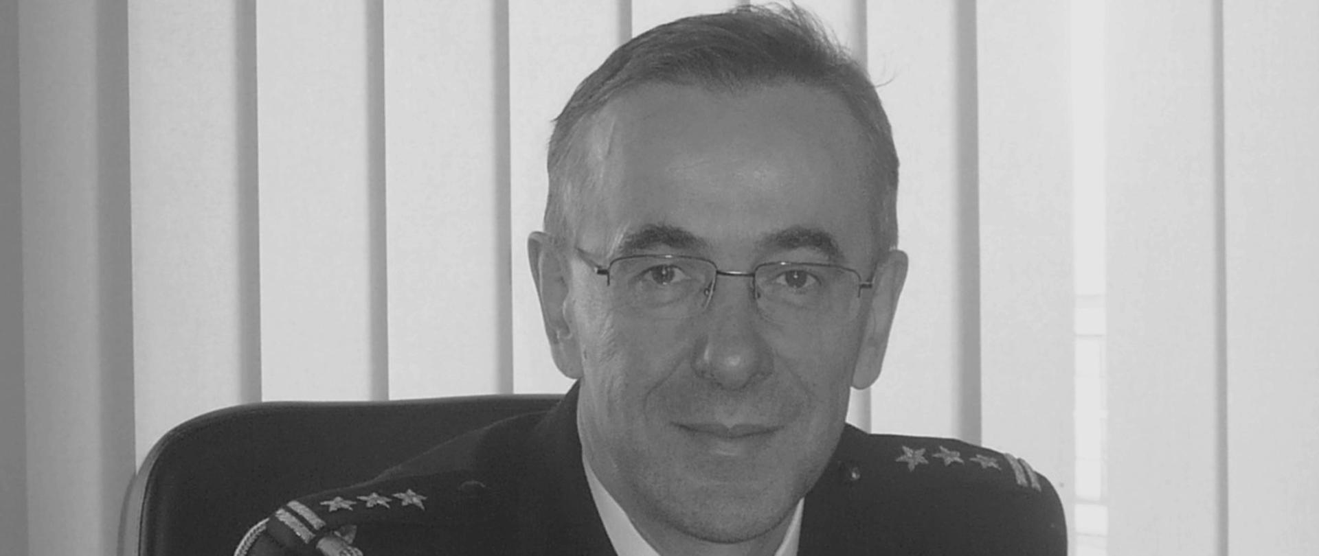 Zdjęcie przedstawia zmarłego wieloletniego komendanta rejonowego i powiatowego tutejszej komendy, st. bryg. w stan. spocz. Andrzeja Stafieja. W pozycji siedzącej przy biurku w mundurze galowym ze sznurem. 