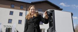 Minister Katarzyna Pełczyńska-Nałęcz stoi przy ładowarce elektrycznej z przodu przed domem.