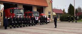 Strażacy stojący w rzędzie, za nimi samochody pożarnicze, obok maszt z powiewającą flagą państwową, 