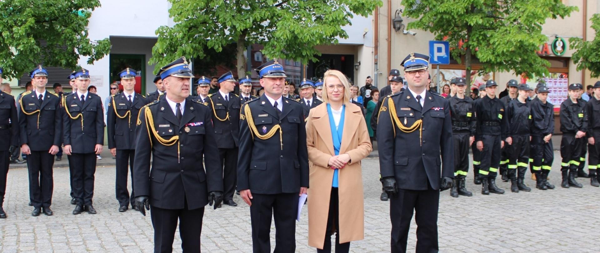 na czerwonym dywanie położonym na kostce brukowej na rynku stoją cztery osoby, trzy z nich to strażacy a jedna to kobieta e jasnym płaszczu i blond włosach. w tle strażacy w szeregu