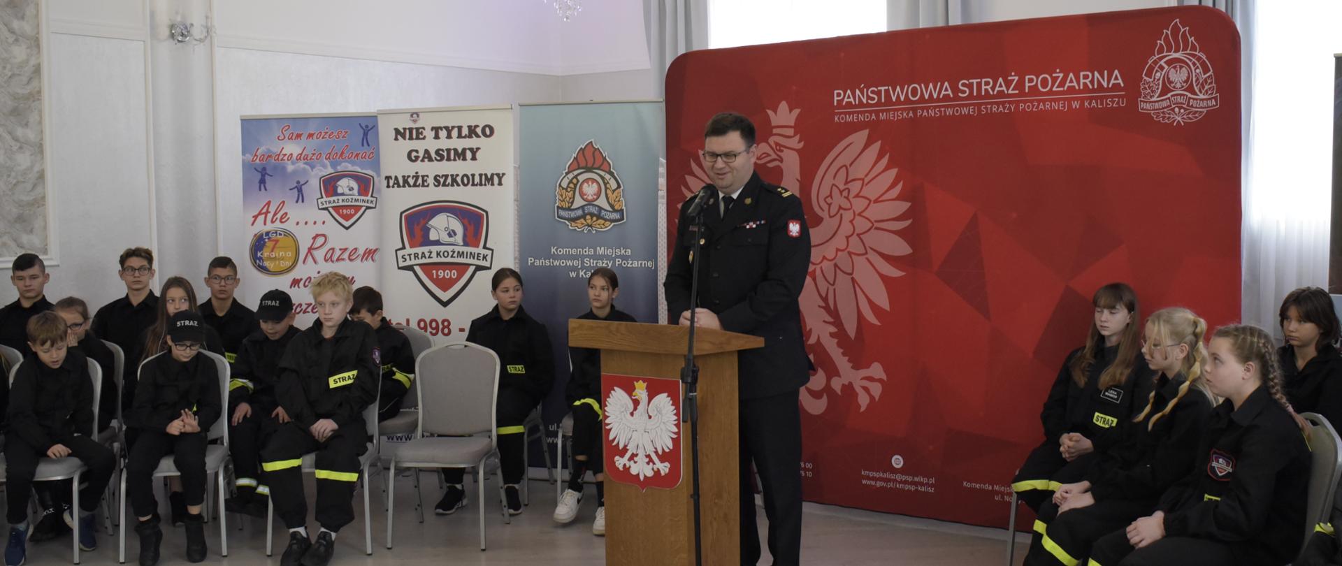 strażak mężczyzna stoi przed mównicą, za nim duża tekstylna ścianka koloru czerwonego na której jest orzeł , logo PSP oraz napis państwowa straż pożarna, po obu stronach na krzesłach siedza dzieci w umundurowaniu strażaka