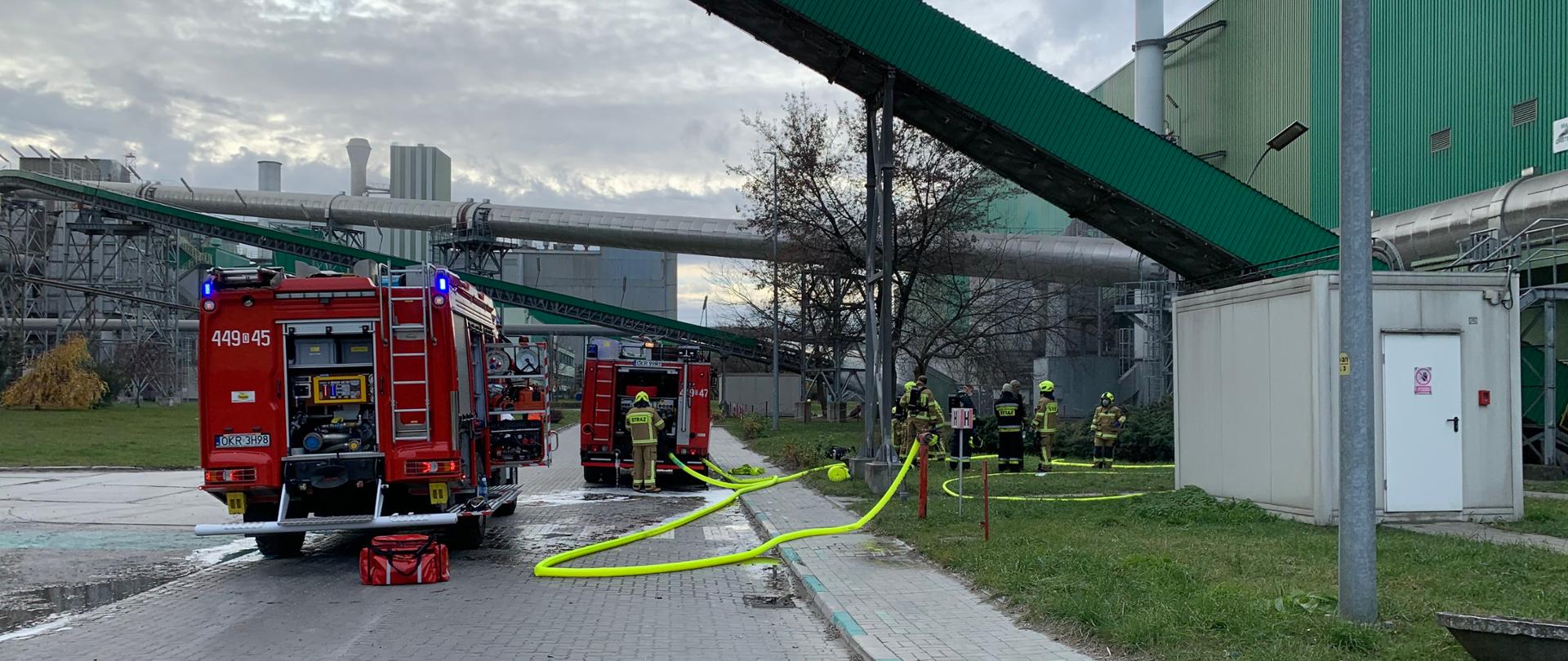 Zdjęcie przedstawia samochody pożarnicze i strażaków podczas gaszenia pożaru w hali magazynowej. Samochody usytuowane po lewej i w środku zdjęcia ustawione tyłem do fotografującego. W prawo w kierunku hali rozwinięte są węże, jeden podpięty do hydrantu zasila jeden z samochodów. Przy samochodzie w środku zdjęcia - bardziej oddalonym stoi kierowca obsługujący pompę. Po prawej stoją strażacy w oczekiwaniu na wejście do hali i obsługujący linie wężowe. Po prawej znajduje się hala magazynowa w kolorze zielonym. W lewo w górę wychodzi z niej obudowany na zielono taśmociąg. W tle inne zabudowania zakładu. 