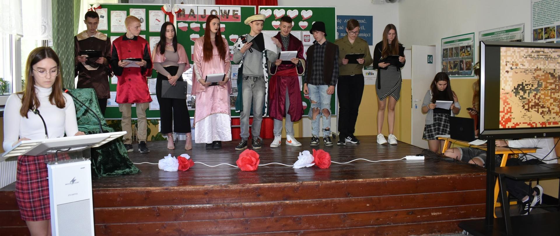 Szerokie ujęcie apelu szkolnego. Grupa uczniów w kostiumach historycznych i strojach galowych stoi na drewnianym podwyższeniu. Po lewej uczennica przy mównicy czyta tekst, po prawej widoczny monitor interaktywny.