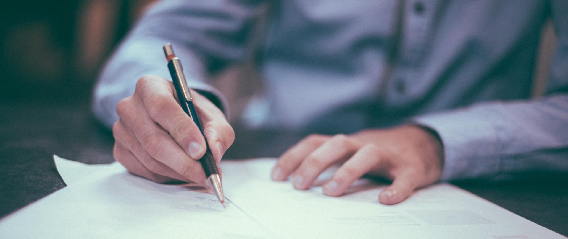 fotografia przedstawia niepełną sylwetkę mężczyzny w niebieskiej koszuli siedzącego przy blacie biurka, podpisującego dokumenty trzymanym w ręce czarnym długopisem