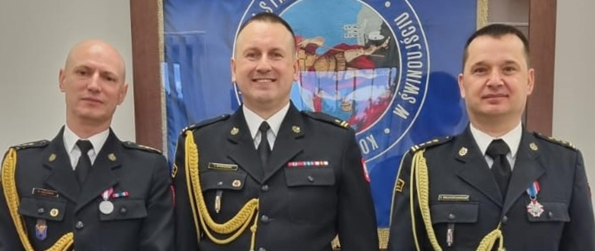 Na zdjęciu widoczni: Komendant Miejski od prawej, Dowódca JRG po środku, Z-ca Komendanta Miejskiego. 3 funkcjonariuszy w umundurowaniu wyjściowym na tle sztandaru KM PSP w Świnoujściu.