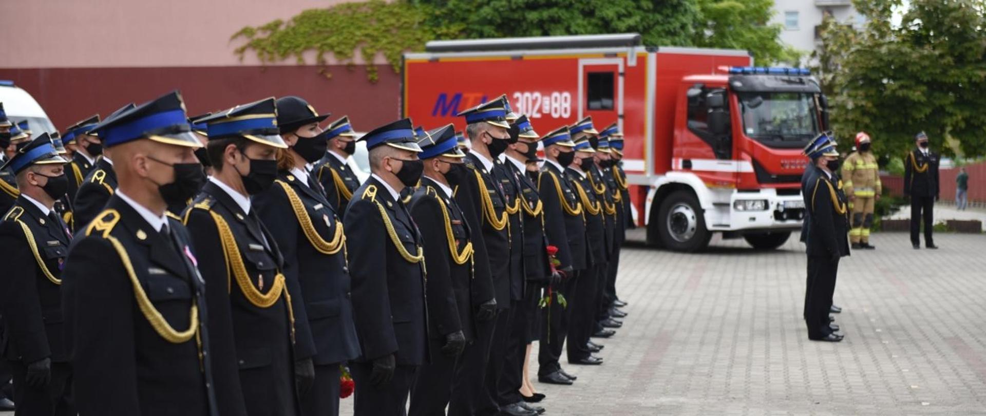 Zdjęcie przedstawia funkcjonariuszy Państwowej Straży Pożarnej ustawionych w szeregach w umundurowaniu galowym oraz czarnych maseczkach. W tle pojazdy pożarnicze. 