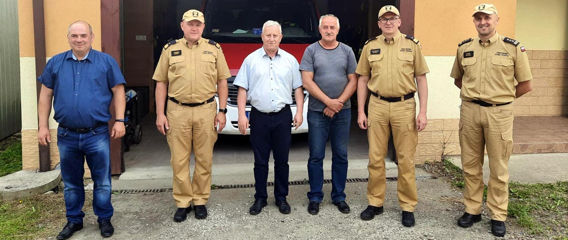 Zdjęcie zrobione na zewnątrz. Przed remizą ochotniczej straży pożarnej stoi sześciu mężczyzn. Trzech z nich to oficerowie Państwowej Straży Pożarnej w mundurach. Za nimi widać samochód ratowniczo-gaśniczy.