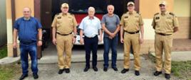 Zdjęcie zrobione na zewnątrz. Przed remizą ochotniczej straży pożarnej stoi sześciu mężczyzn. Trzech z nich to oficerowie Państwowej Straży Pożarnej w mundurach. Za nimi widać samochód ratowniczo-gaśniczy.