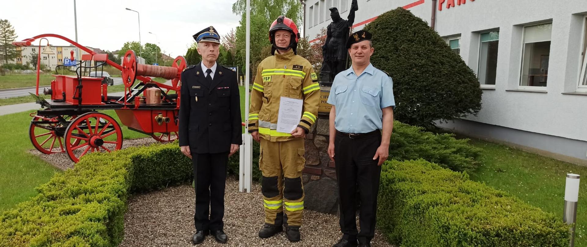Zdjęcie przedstawia trzech strażaków stojących przy pomniku św. Floriana jeden z nich ubrany jest na bojowo, dwóch w mundurach wyjściowych.