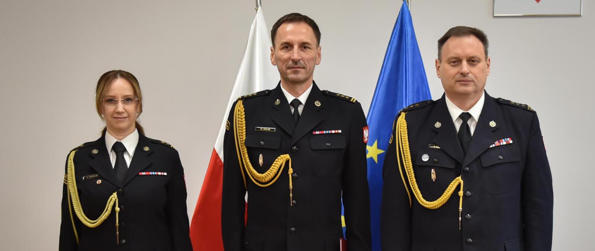 troje strażaków pozujących do zdjęcia w gabinecie, za nimi flagi polski i unii europejskiej