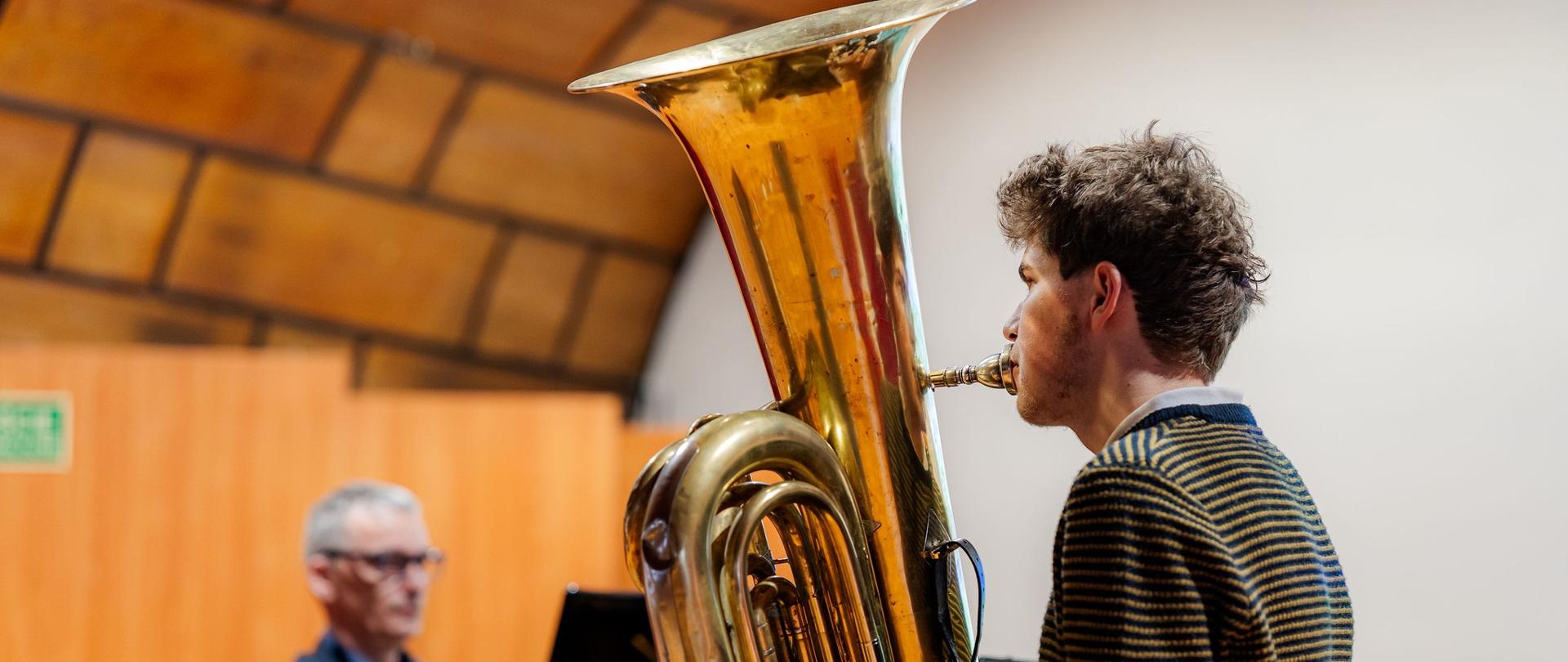 Młody muzyk w pasiastym swetrze gra na tubie. Instrument jest duży i błyszczący. W tle, przy czarnym fortepianie, siedzi akompaniator w okularach.