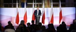 Widok z tyłu na zaciemnioną wypełnioną ludźmi salę, przed nimi na oświetlonym podwyższeniu stoi przy mównicy minister Czarnek i mówi do mikrofonu, za nim rząd sześciu polskich flag.