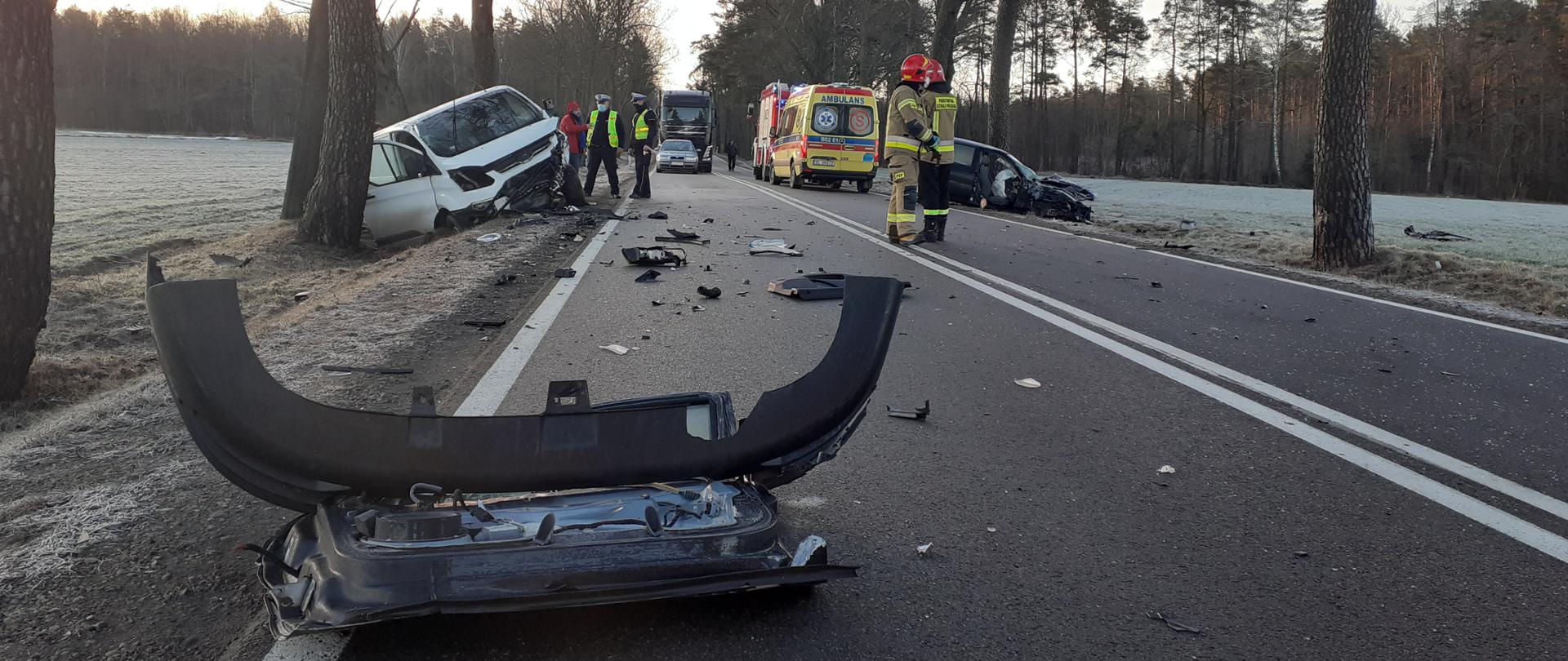 Na zdjęciu widać pas drogowy, na którym na pierwszym planie leży urwany zderzak pojazdu. W dalszej części w rowach po przeciwnych stronach widać dwa rozbite pojazdy. Na drodze stoi karetka pogotowia i samochód pożarniczy dwóch strażaków i 2 policjantów.