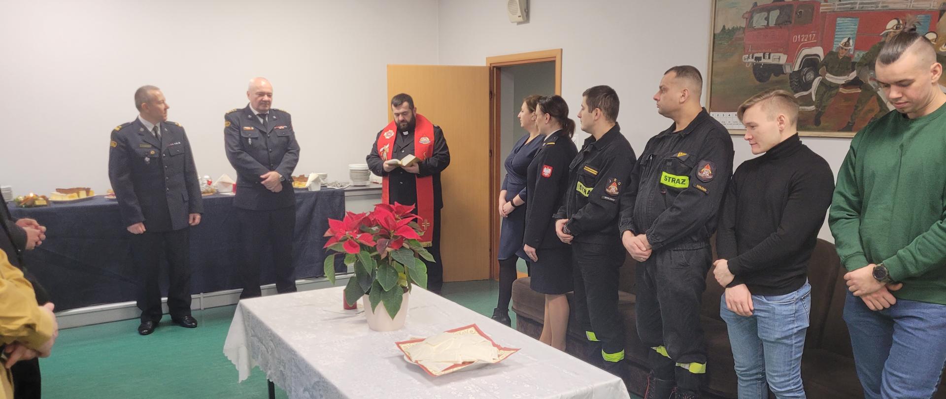 Zdjęcia przedstawiają strażaków z KP PSP Wołomin podczas spotkania wigilijnego w komendzie. Na zdjęciach widoczni są funkcjonariusze w mundurach wyjściowych oraz koszarowych. Pośród strażaków jest również ksiądz kapelan. Strażacy stoją wokoło stołu nakrytego białym obrusem na którym stoi opłatek. na innych zdjęciach widać strażaków dzielących się opłatkiem i skłądających sobie życzeniaz.